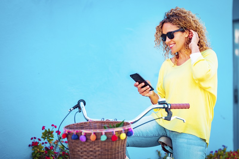 Portrait of attractive adult middle age female smiling at mobile phone sitting on a bike against a blue wall in background. Woman people enjoy technology connection outdoor